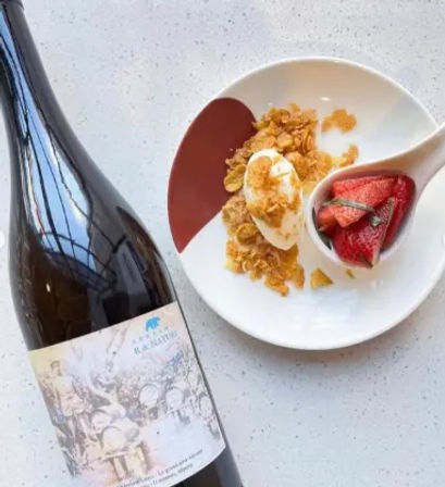 Dark wine bottle with decorative label beside a white plate on a speckled countertop, showing a dollop of cream topped with crunchy cornflakes and a small spoon of sliced strawberries — playful wine-and-dessert pairing.