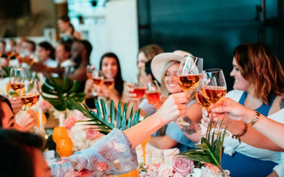 Smiling group toasting with rosé wine glasses at a festive dining table decorated with flowers and palm leaves, a cheerful celebration.