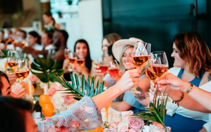 Smiling group toasting with rosé wine glasses at a festive dining table decorated with flowers and palm leaves, a cheerful celebration.