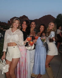 Five friends in summer outfits raising colorful cocktails on a rooftop terrace at dusk with red rock desert hills and a clear sky in the background.