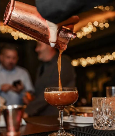 Copper cocktail shaker pouring an amber cocktail into a coupe glass at a dimly lit bar with warm bokeh string lights in the background