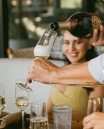 Server pouring sparkling champagne into a flute at a restaurant table, smiling guest in the background and multiple glasses on the table.
