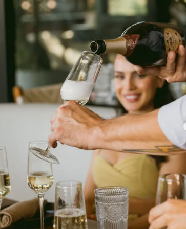 Server pouring sparkling champagne into a flute at a restaurant table, smiling guest in the background and multiple glasses on the table.