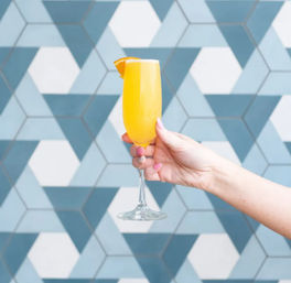 Hand with pink nails holding a bright orange mimosa in a champagne flute garnished with an orange slice, set against a blue geometric tile wall.