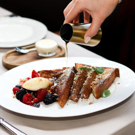 Hand pouring maple syrup from a small metal pitcher over sliced French toast dusted with powdered sugar and chopped nuts, served with mixed berries and a scoop of cream on a white plate at brunch.