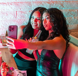Two friends in a neon-lit lounge booth posing for a selfie — one holds a cocktail, the other a bottled drink, with a textured brick-wall backdrop.