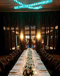 Intimate, dimly lit upscale restaurant dining room with a long white-clothed banquet table lined with leather chairs, candlelit glassware and a white floral centerpiece runner beneath a modern turquoise LED chandelier.