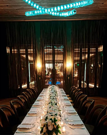 Intimate, dimly lit upscale restaurant dining room with a long white-clothed banquet table lined with leather chairs, candlelit glassware and a white floral centerpiece runner beneath a modern turquoise LED chandelier.