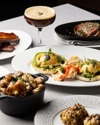 Upscale brunch spread with lobster eggs Benedict topped with avocado and hollandaise, crispy bacon, seared steak, roasted potatoes in a cast-iron pot, and an espresso martini on a white table.