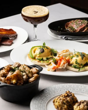 Upscale brunch spread with lobster eggs Benedict topped with avocado and hollandaise, crispy bacon, seared steak, roasted potatoes in a cast-iron pot, and an espresso martini on a white table.