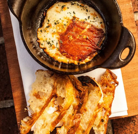 Oozy baked cheese dip in a small cast-iron skillet topped with crispy cured ham and herbs, served with toasted sourdough slices on a wooden board — warm appetizer.