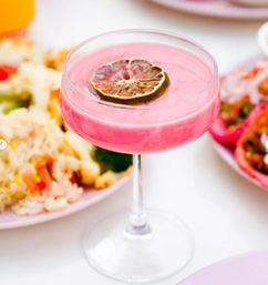 Bright pink creamy cocktail in a coupe glass topped with a dried lime slice, set on a white table beside colorful plates of rice and tacos — vibrant summer brunch drink.