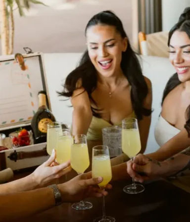 Smiling friends toasting with yellow mimosas in flutes at a lively indoor brunch, champagne bottle and bowl of strawberries on the table.