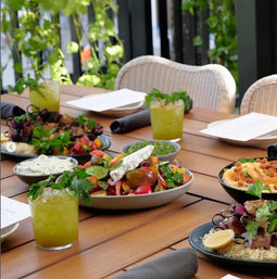 Al fresco patio spread on a wooden table with colorful Mediterranean mezze — heirloom tomato and feta salad, hummus, grilled skewers over rice, herb‑garnished green cocktails, plates and wicker chairs beside a leafy backdrop.