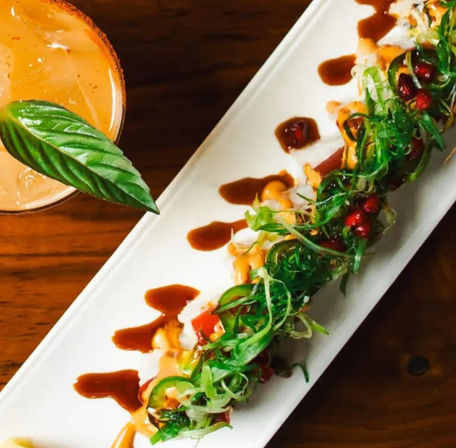 Vibrant sushi roll on a rectangular white plate topped with seaweed salad, sliced jalapeño, pomegranate seeds and drizzled spicy mayo and eel sauce, paired with a basil‑garnished cocktail on a wooden table.