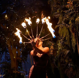 Fire performer holding a fan of flaming torches at night in a lush garden setting, flames arcing in a semicircle against twinkling lights.
