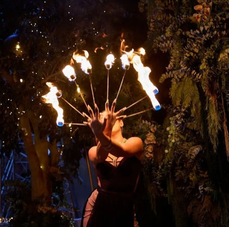 Fire performer holding a fan of flaming torches at night in a lush garden setting, flames arcing in a semicircle against twinkling lights.