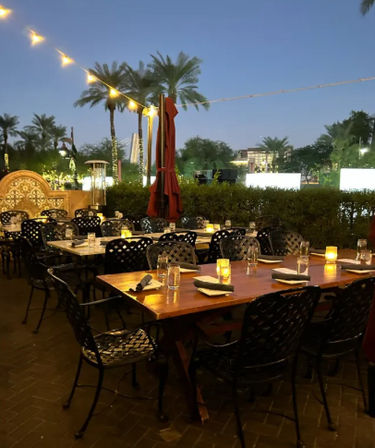 Al fresco dinner patio at dusk with wooden tables set with napkins and glasses, candlelit votives, string lights and palm trees overhead.
