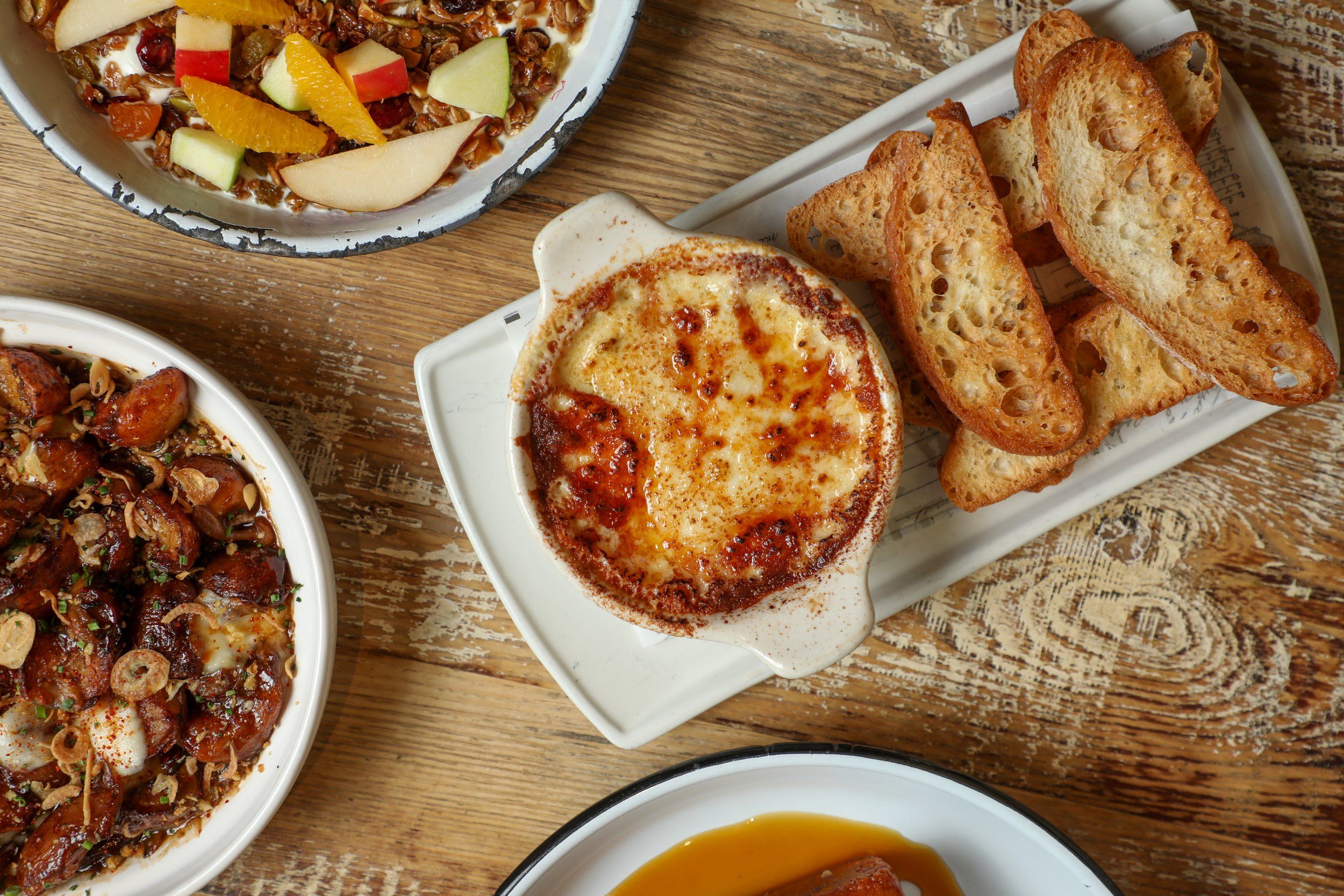 Top-down brunch spread on a rustic wooden table: bubbling baked cheese dip with toasted crostini, fruit-topped granola bowl, and roasted potatoes.