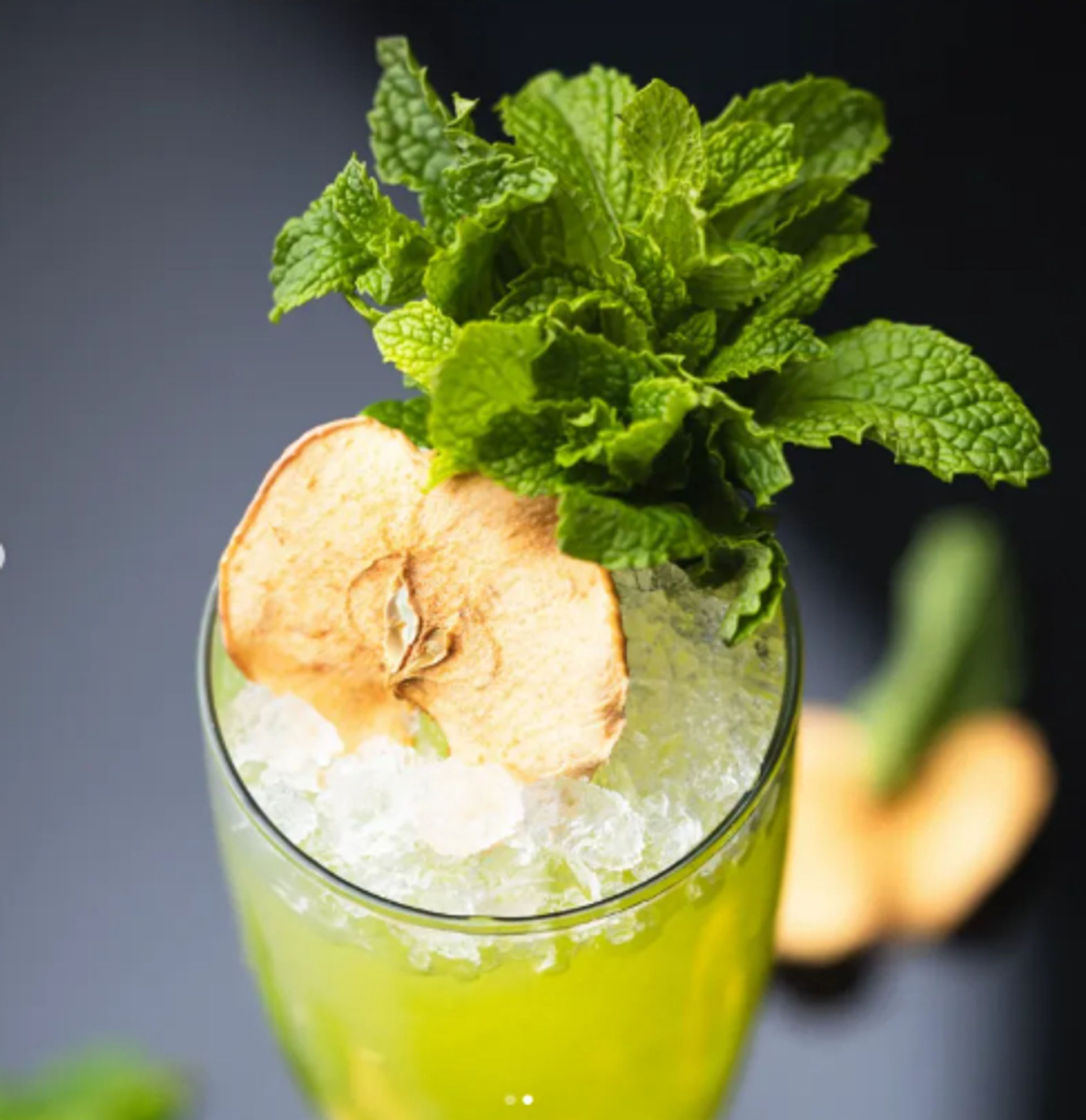 Bright green mint cocktail in a tall glass filled with crushed ice, topped with a fresh mint sprig and a dried apple slice garnish