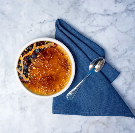 Top-down view of a golden caramelized crème brûlée in a white ramekin, topped with blueberries, candied orange peel and chopped nuts, set on a blue napkin with a silver spoon on a marble countertop.