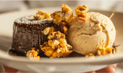 Close-up of a gooey chocolate lava cake with glossy chocolate sauce, a scoop of caramel ice cream, and crunchy caramel popcorn on a white dessert plate