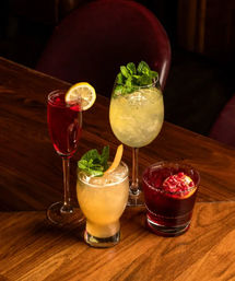 Four colorful craft cocktails on a wooden bar table: a tall red flute with lemon slice, a large minty yellow‑green spritz, a peachy short glass with mint and peel, and a deep red pomegranate cocktail.