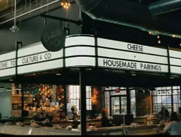 Industrial market hall interior with a bright marquee sign reading 'CHEESE' and 'HOUSEMADE PAIRINGS' above an artisan cheese counter and seated patrons.