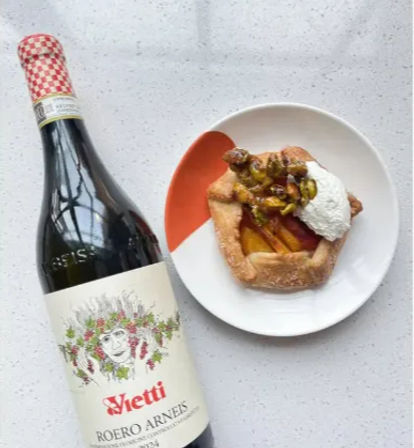 Bottle of white wine beside a rustic peach galette topped with whipped cream and pistachios on a white plate — wine and dessert pairing on a speckled countertop.