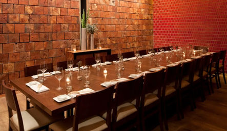 Long wooden dining table set for a private dinner in a cozy, modern-rustic restaurant room with rows of chairs, wine glasses, napkins, lit votive candles and textured wood and red-tile walls