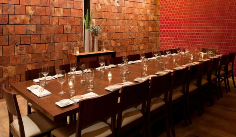 Long wooden dining table set for a private dinner in a cozy, modern-rustic restaurant room with rows of chairs, wine glasses, napkins, lit votive candles and textured wood and red-tile walls