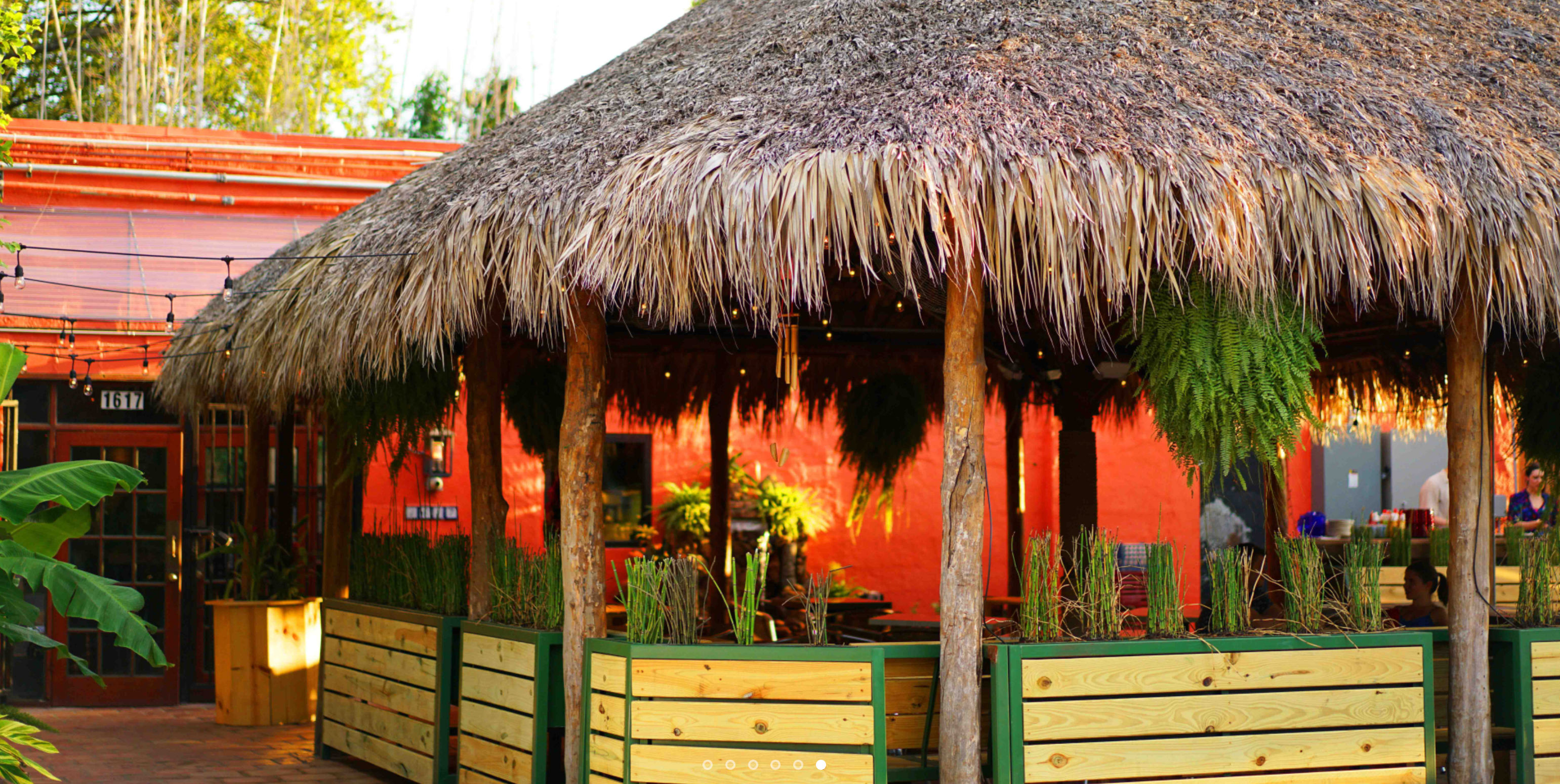 Thatched-roof tiki patio with hanging ferns, string lights and wooden planters in a colorful tropical outdoor dining courtyard