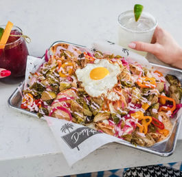 Brunch tray of loaded breakfast nachos topped with a sunny-side-up egg, pickled red onions, sliced bell peppers, roasted Brussels sprouts and pink crema, served on a metal tray with a margarita and a red cocktail nearby