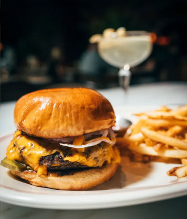 Glossy brioche cheeseburger with melted cheddar, pickles and sliced onions on a white plate, paired with crispy French fries and a blurred cocktail on a restaurant table