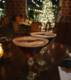 Two espresso martini cocktails in vintage coupe glasses on a dark wood table at a cozy restaurant bar, blurred string lights and a lit holiday tree in the background.