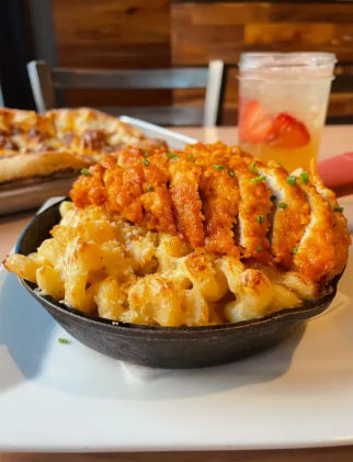 Crispy buffalo chicken slices atop cheesy baked macaroni and cheese served in a cast-iron skillet on a casual restaurant table, with a pizza slice and strawberry-infused drink blurred in the background