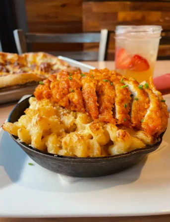 Crispy buffalo chicken slices atop cheesy baked macaroni and cheese served in a cast-iron skillet on a casual restaurant table, with a pizza slice and strawberry-infused drink blurred in the background