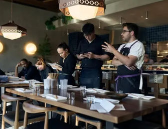 Restaurant team huddle by a long wooden communal table set for service, chefs and servers checking notes and phones with an open kitchen and pendant lights in the background.