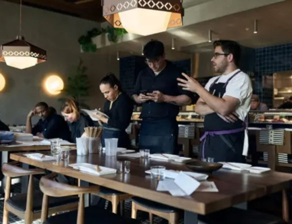 Restaurant team huddle by a long wooden communal table set for service, chefs and servers checking notes and phones with an open kitchen and pendant lights in the background.