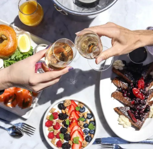 Sunlit brunch spread with two hands clinking champagne glasses over a marble table — smoked salmon bagel, glass of orange juice, granola and mixed berry bowl, and berry-topped French toast.