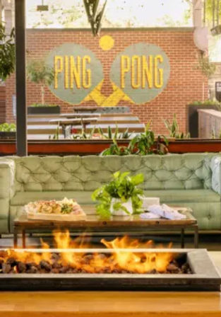 Urban outdoor patio lounge with a rectangular fire pit, tufted sofa, wooden coffee table with plants, and a brick wall mural featuring colorful ping-pong paddles