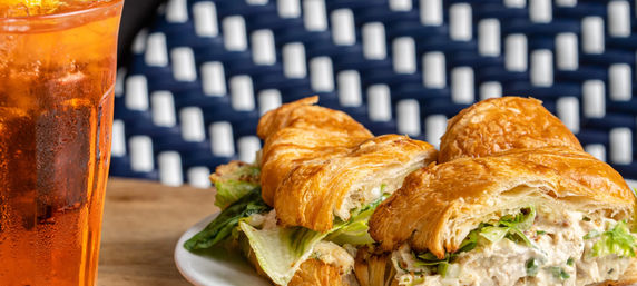 Golden flaky croissant chicken-salad sandwich with crisp lettuce on a white plate beside a frosty amber iced tea, set on a wooden table with a blue bistro chair backdrop