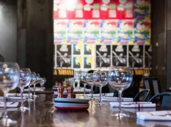 Long communal wooden table set with empty wine glasses, napkins and salt-and-pepper shakers in a trendy urban bistro with a colorful postered wall