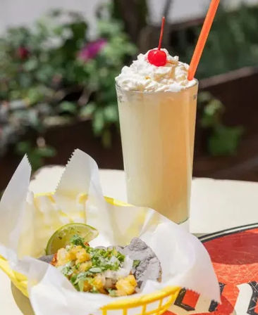 Blue-corn seafood taco with pineapple salsa, cilantro and lime in a yellow paper-lined basket beside a tall creamy vanilla milkshake topped with whipped cream and a cherry, casual Mexican taqueria patio scene