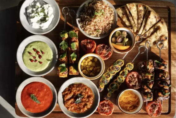 Colorful appetizer platter on a wooden board with assorted dips, grilled skewers, flatbread wedges, rice, roasted tomatoes and pickled vegetables.