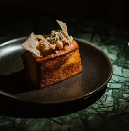 Gourmet appetizer — golden toasted brioche cube topped with creamy herbed relish and crisp cracker shards on a black plate over a green marble table, fine dining presentation.