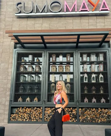 Woman posing by a modern restaurant facade with metal-grid shelves of decorative lanterns and stacked firewood, wearing a crop top and red crossbody bag.