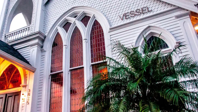 White Victorian-style clapboard building with tall Gothic arched stained-glass windows, tropical palm in front and warm entry lights at dusk.