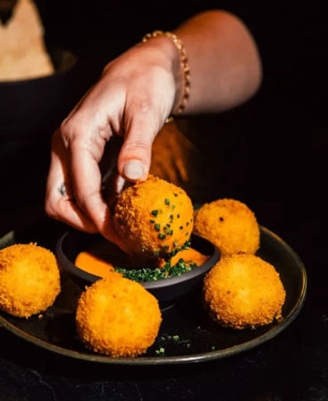 Hand picking a golden, crispy arancini cheese croquette from a dark plate to dip into creamy orange sauce garnished with chopped chives — restaurant appetizer.