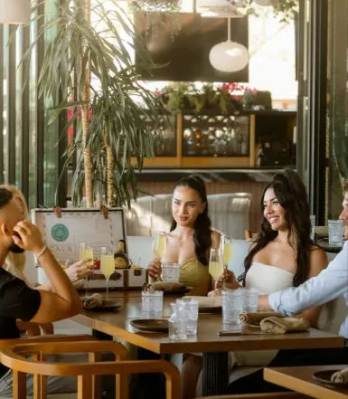 Four friends enjoying a sunny brunch at a modern restaurant patio, smiling and toasting with mimosa glasses around a wooden table surrounded by plants and pendant lights.